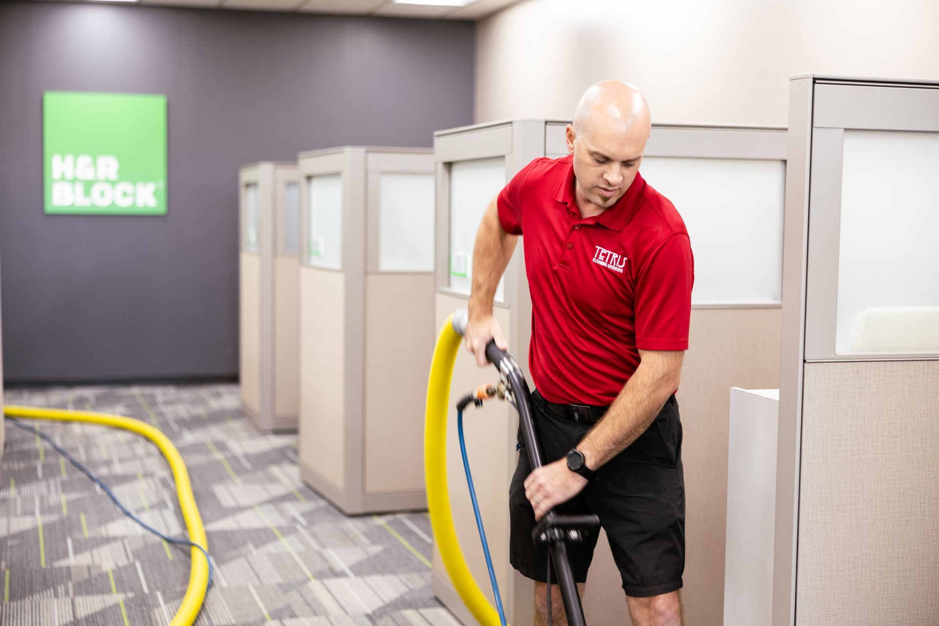 Man vacuuming a carpeted office hallway beside cubicles and a green M&R Block sign