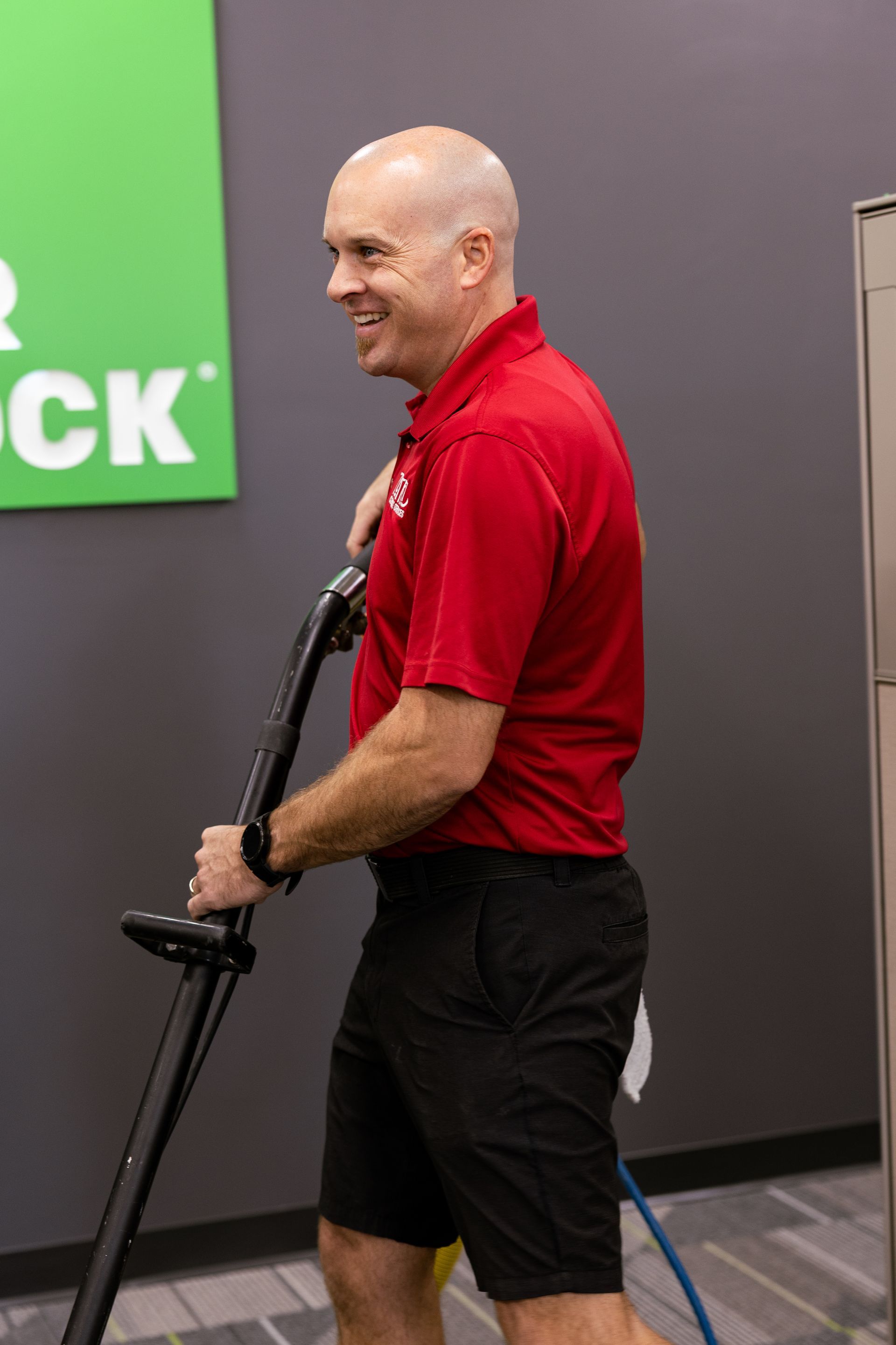 Man in a red shirt and black shorts holding a golf club indoors near a green sign.