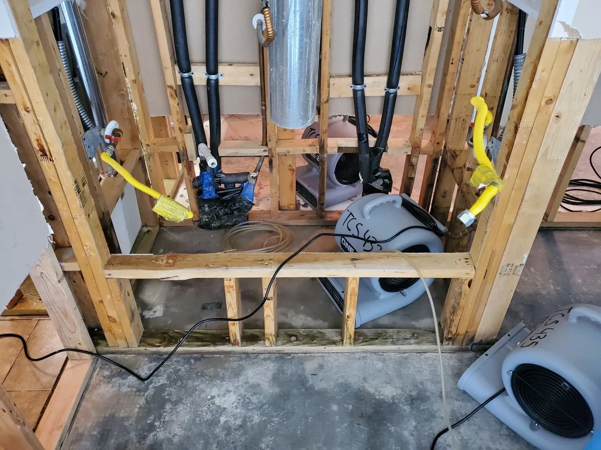 Open wall framing with exposed plumbing, yellow straps, and a gray fan on the floor.