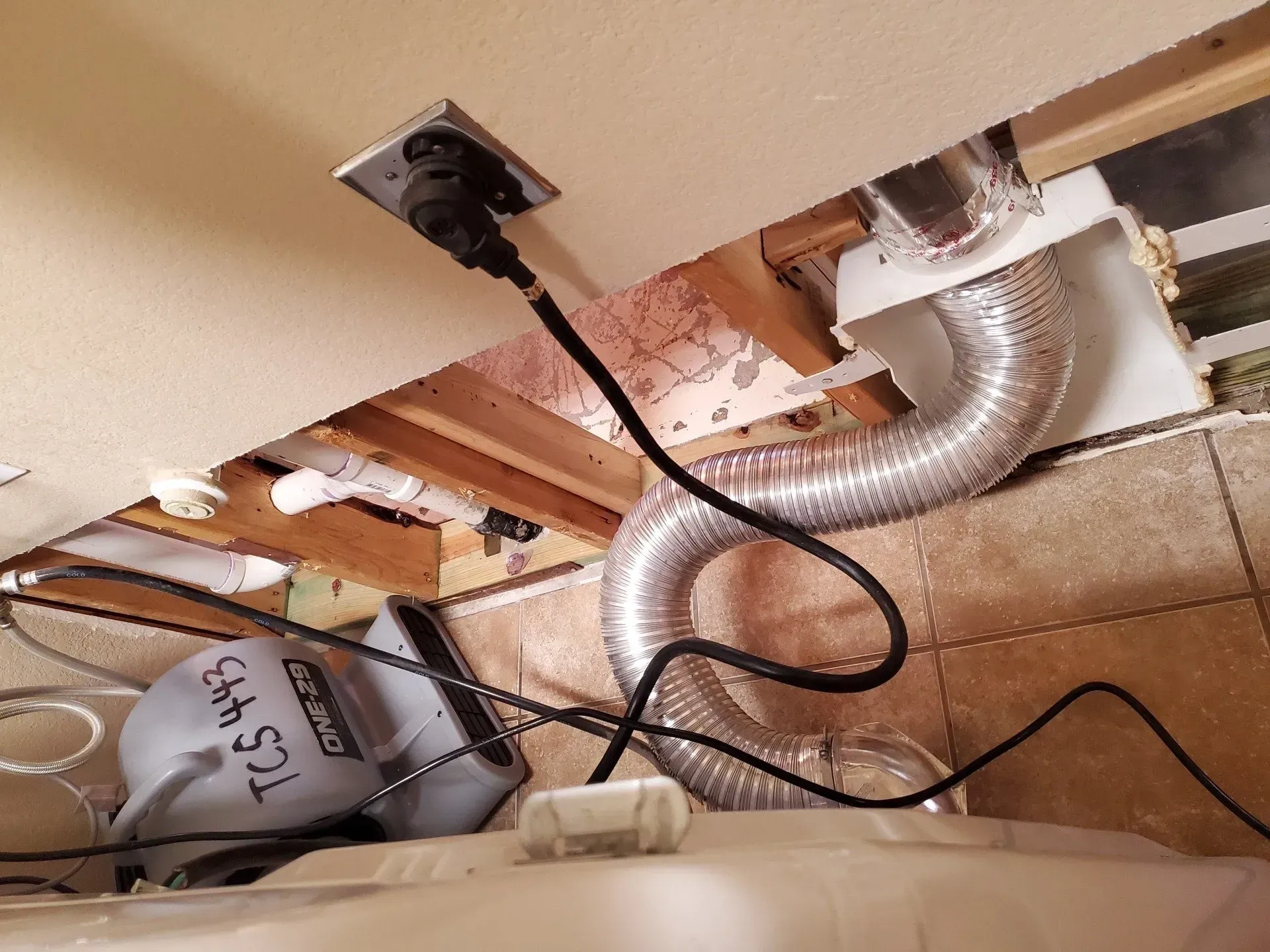 Under-sink cabinet with exposed plumbing, silver ducting, black cables, and a covered bucket on the floor