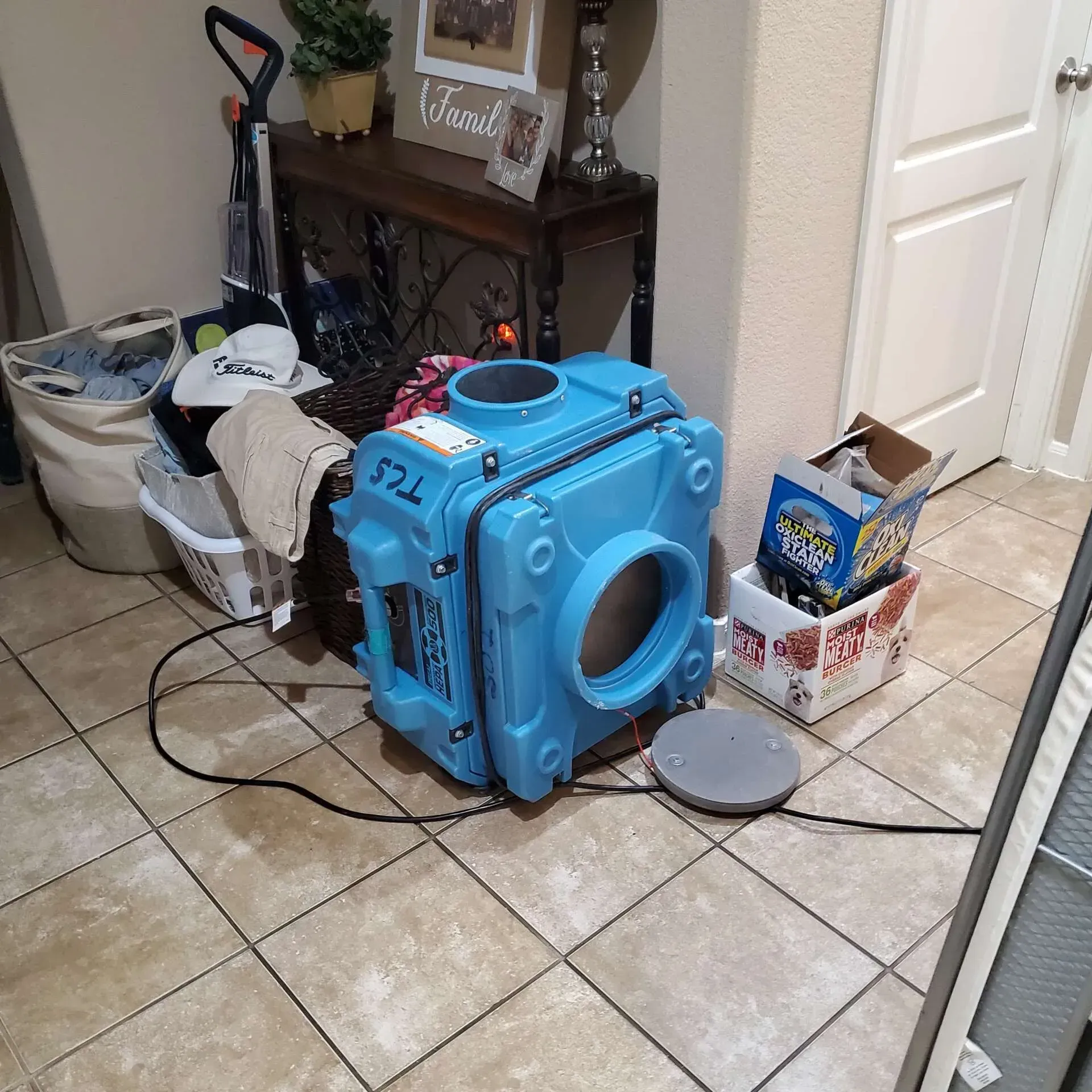 Blue HVAC duct fan on a tiled floor beside a side table, laundry basket, and open box in a hallway.