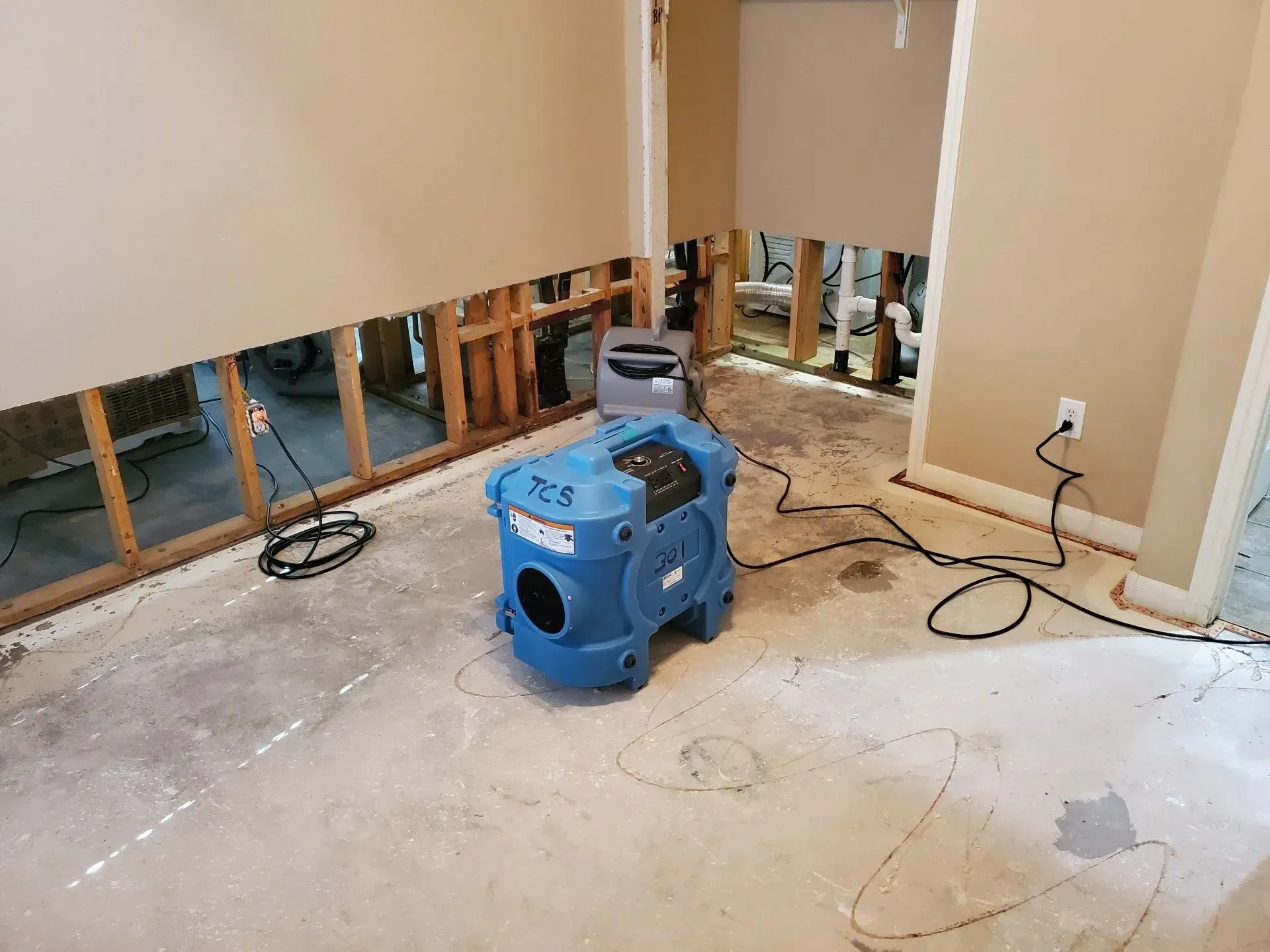 Basement with water-damaged walls, exposed studs, and a blue dehumidifier on the concrete floor.