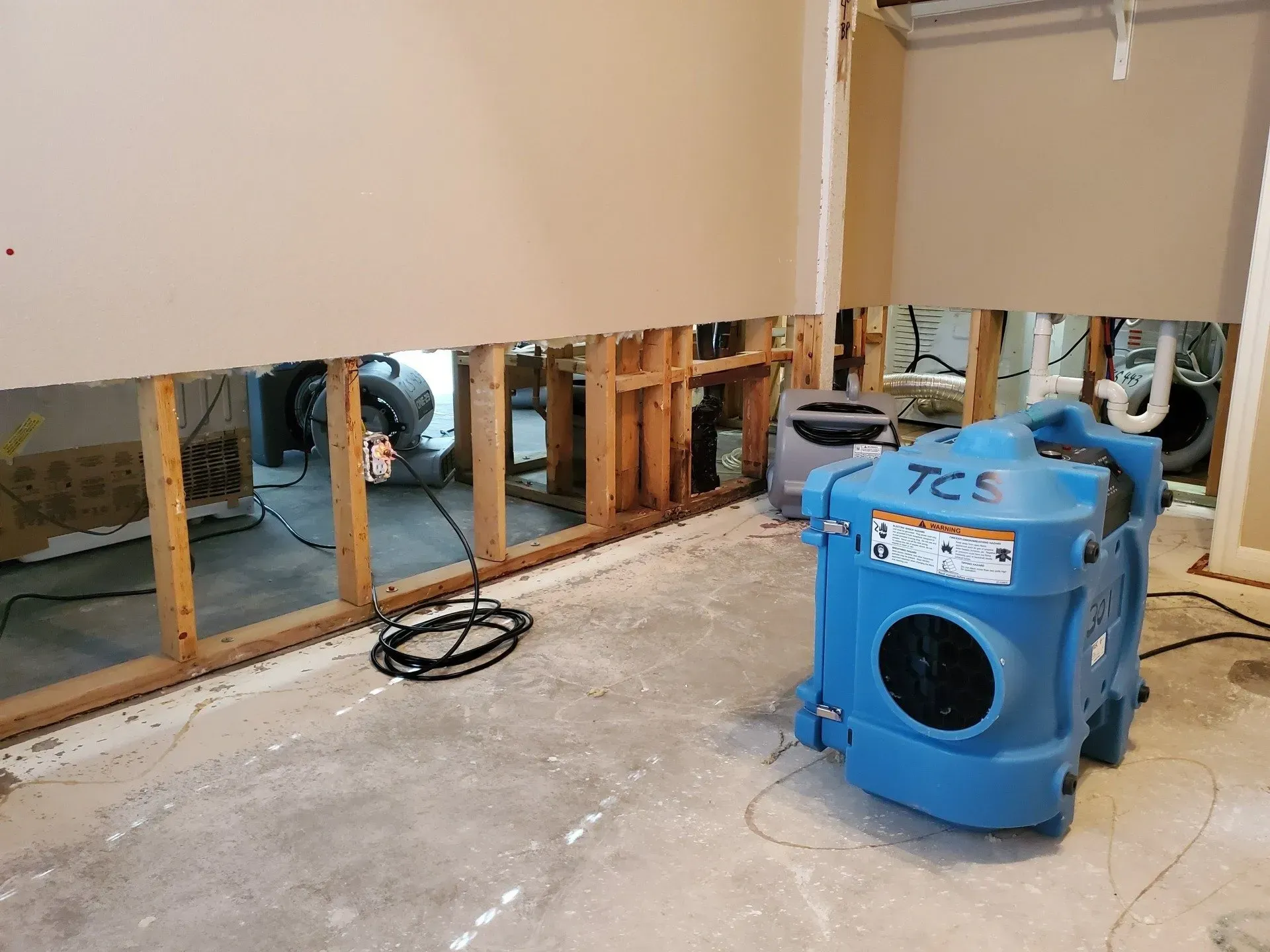 Partially demolished interior with exposed studs, debris, and a blue dehumidifier or blower on the floor