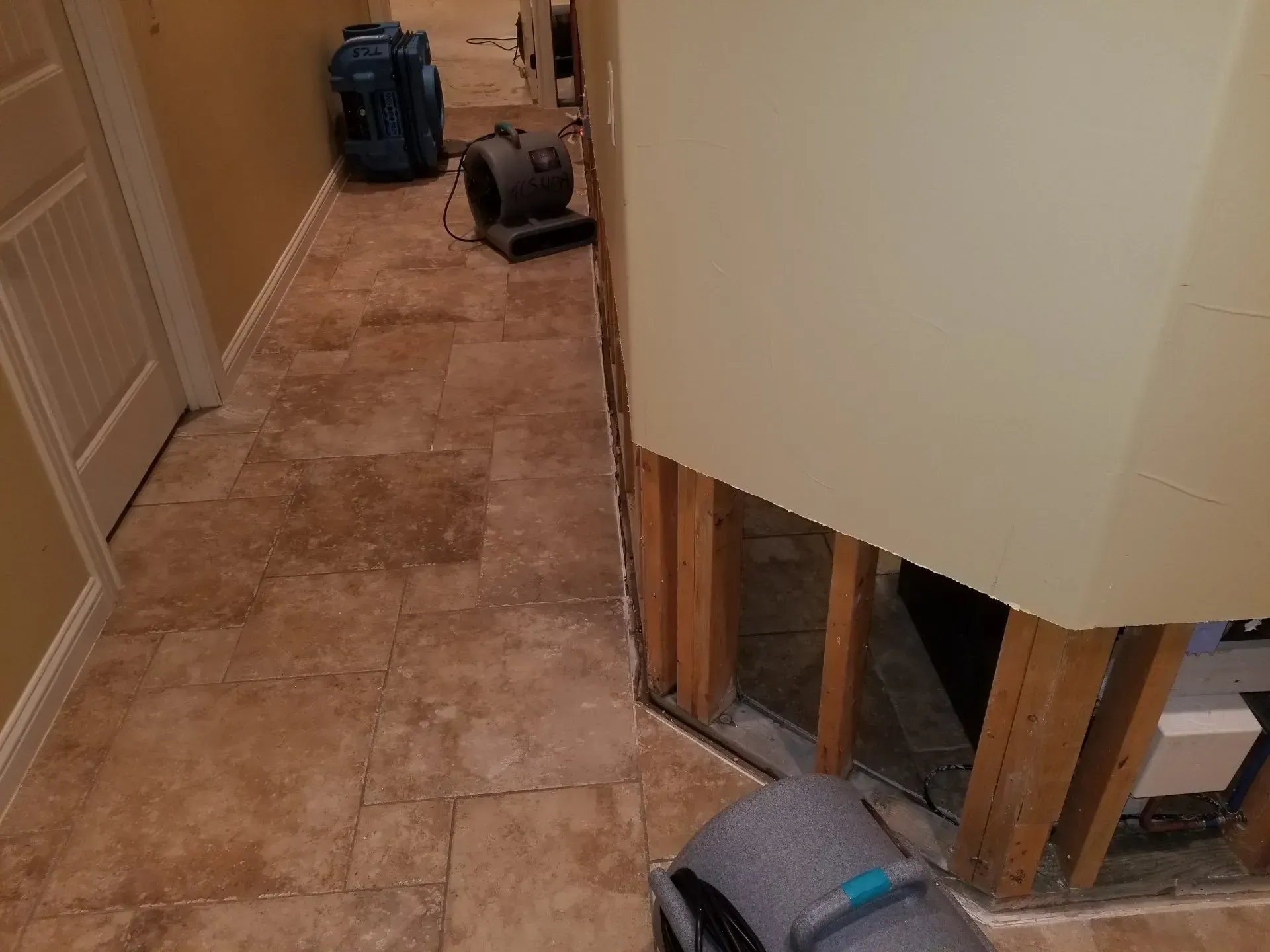 Narrow hallway with tan tile floor, backpack and vacuum at the far end, and exposed wall framing on the right.