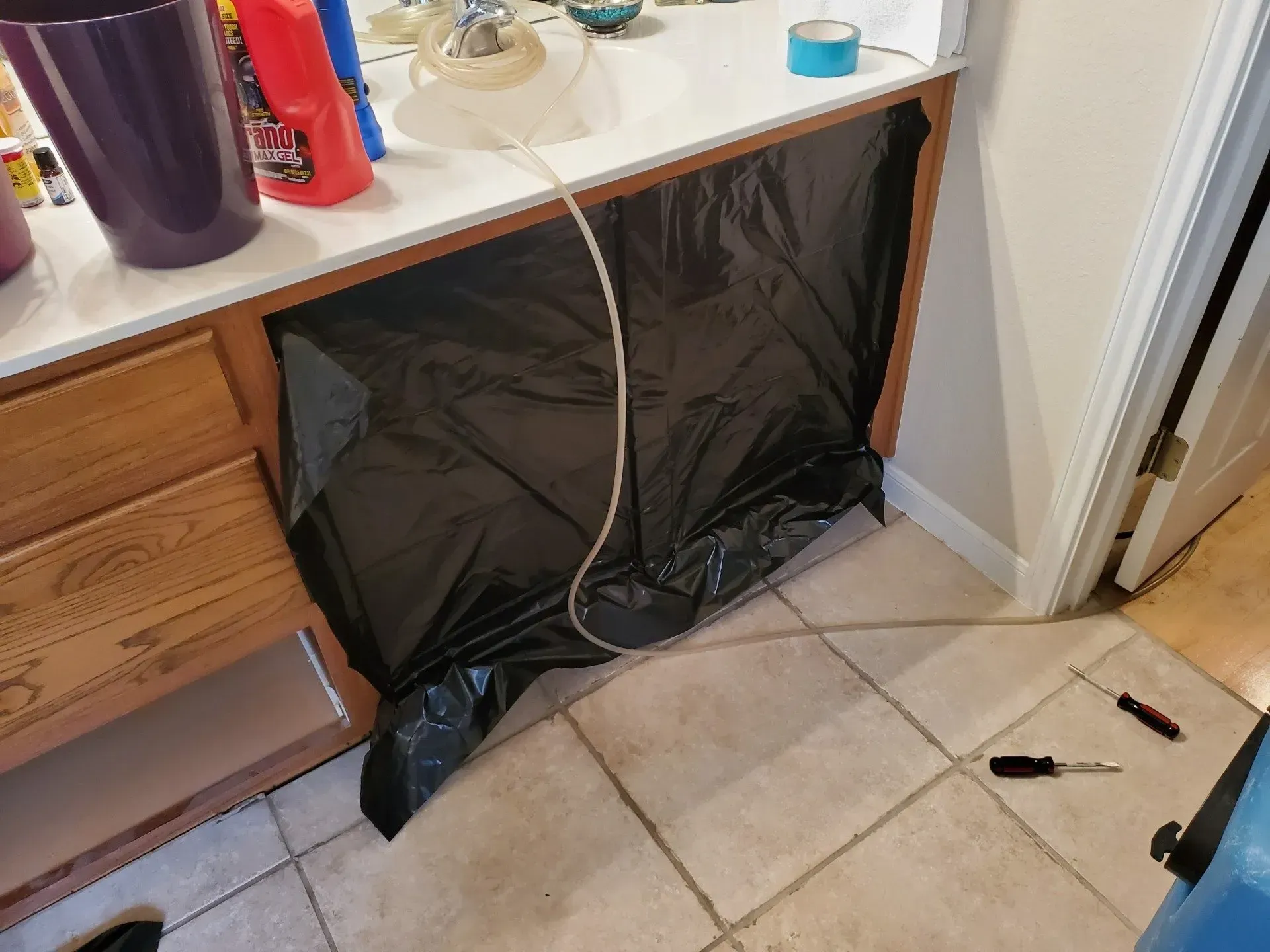 Open cabinet under bathroom sink covered with a black plastic barrier and a hose or cable hanging down.