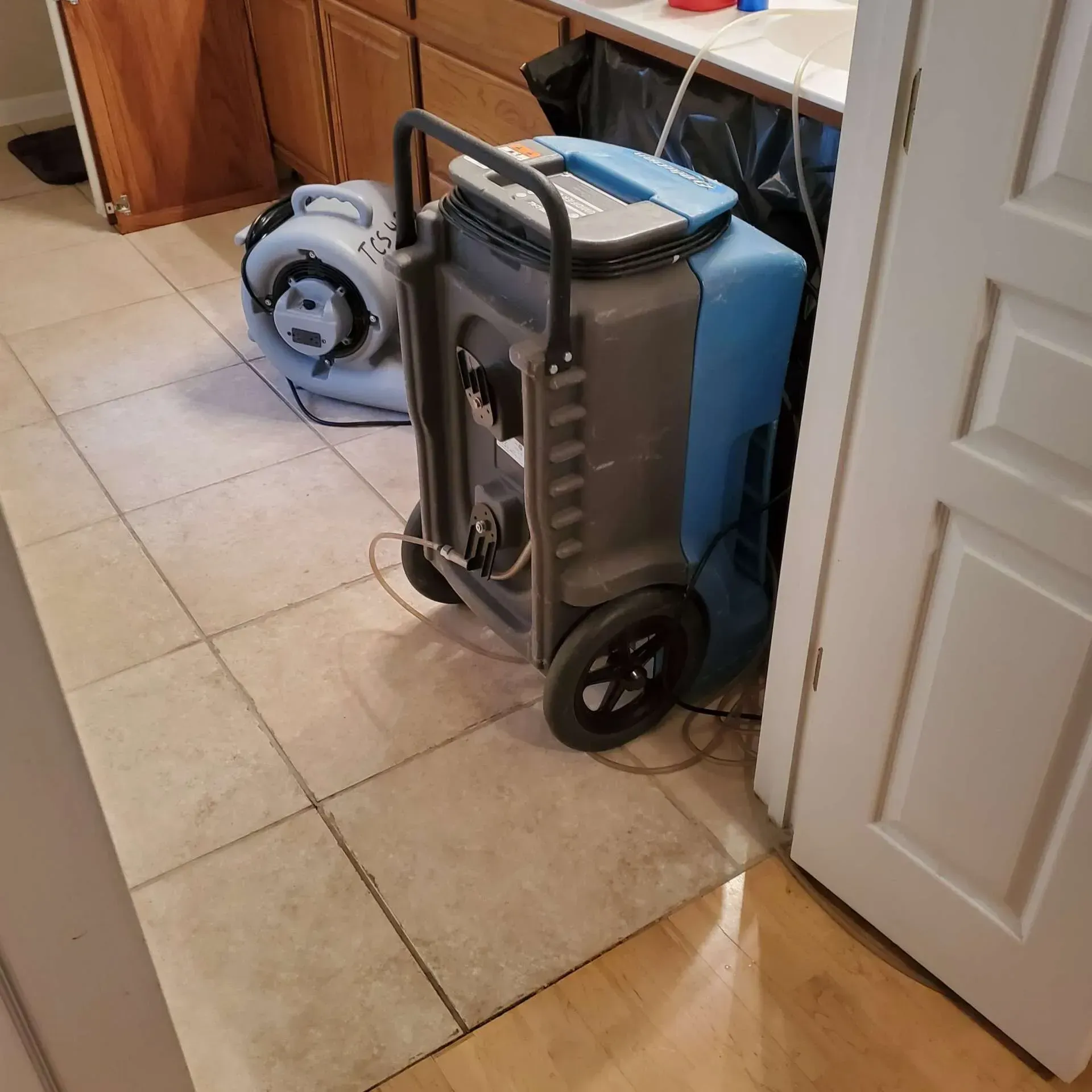 Folded gray hand truck and blue cooler beside a doorway on a tiled floor.