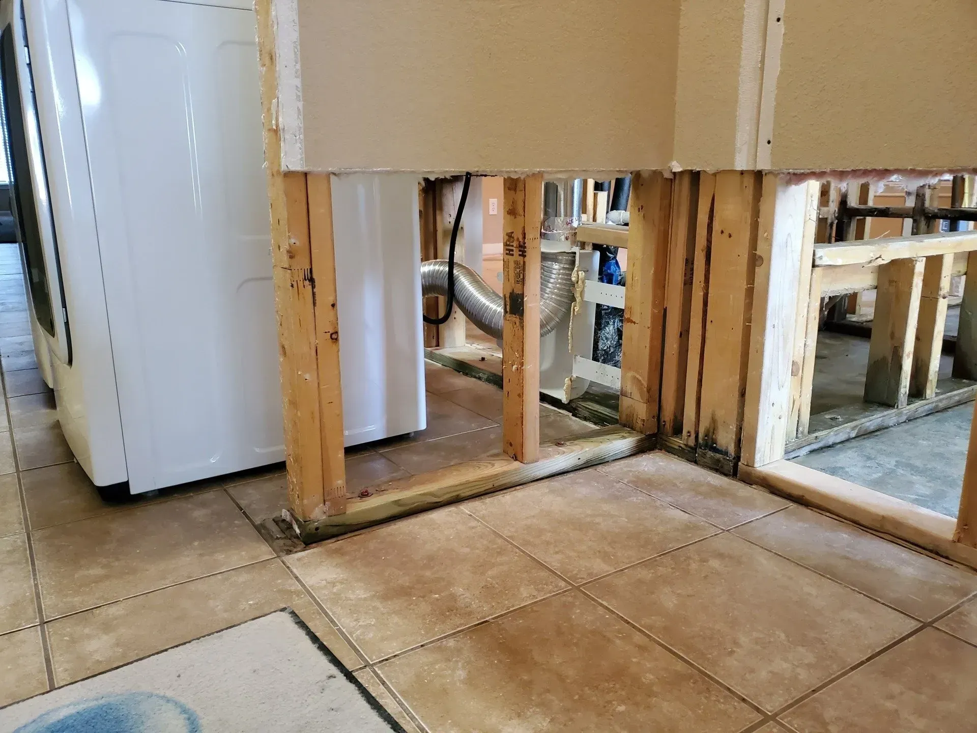 Damaged wall framing and baseboard exposed beside a white refrigerator on a tiled kitchen floor