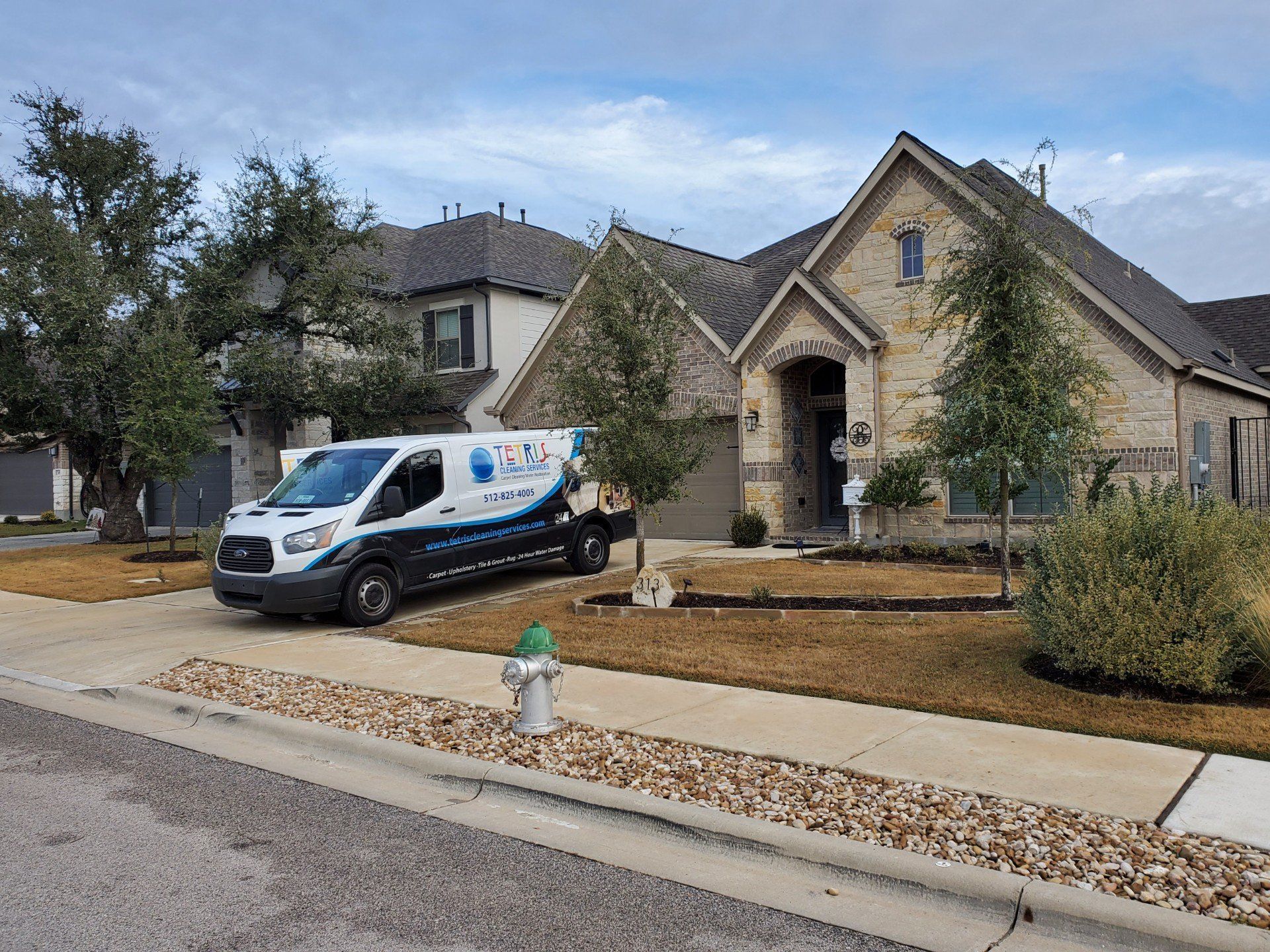 White service van parked outside a tan stone house with a front yard and sidewalk trees.