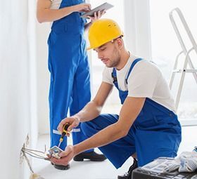 Two electricians installing an outlet. One kneels, wearing a hard hat, while another holds a tablet.