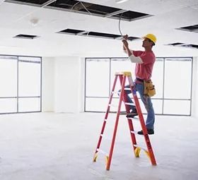 Electrician on a ladder, working on the ceiling wiring in a large, empty room with windows.