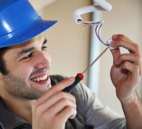 Electrician smiles while wiring an outlet, wearing a blue hard hat.