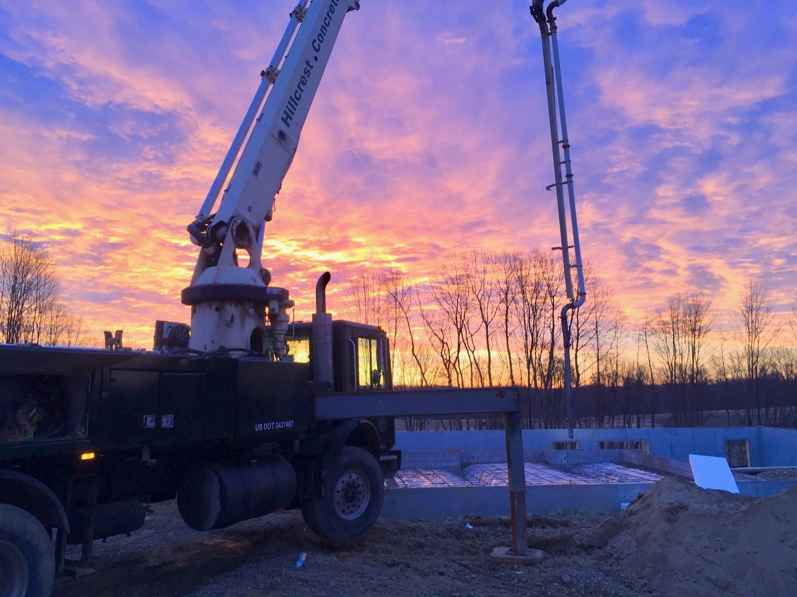 Concrete pump truck pouring concrete into a foundation at sunset. The sky is filled with pink and orange hues.