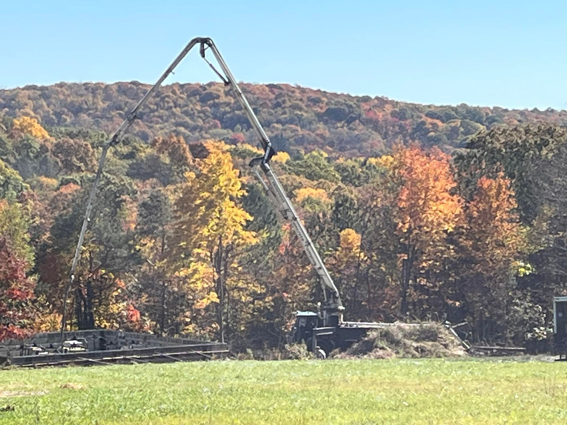 Concrete pump truck in a field, autumn foliage in the background, pumping concrete over a structure.