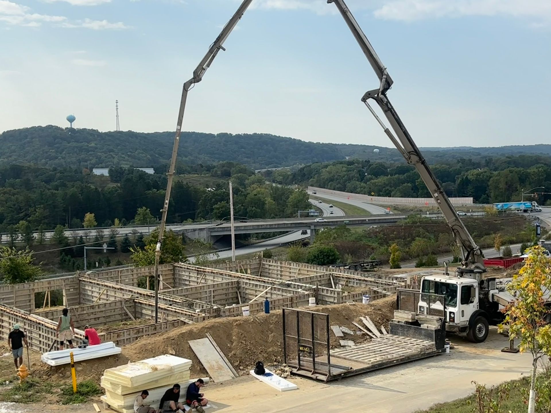 Construction site with concrete pump truck pouring concrete into wooden forms for a building foundation, green landscape in the background.