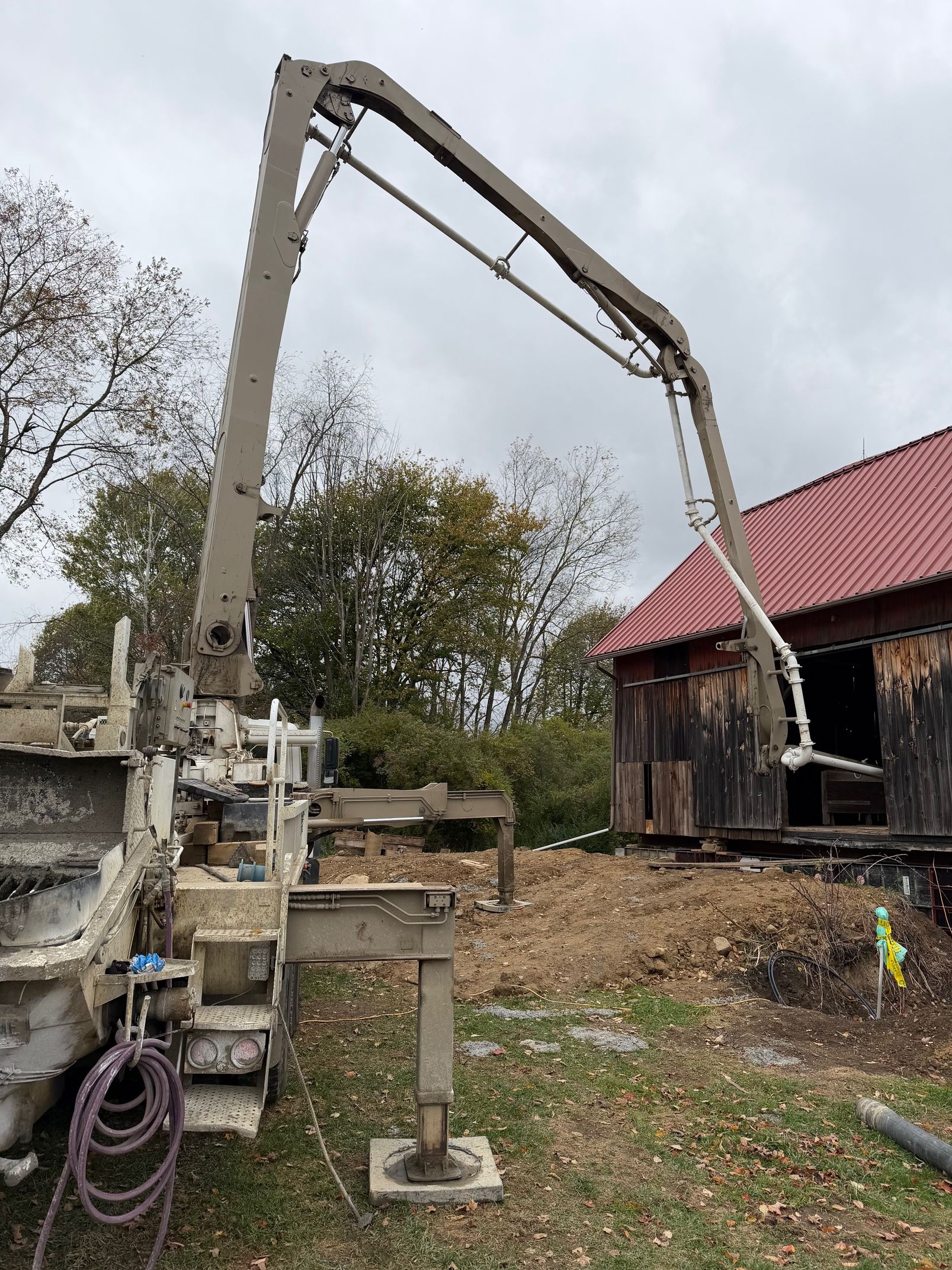 Concrete pump truck with extended boom, pouring concrete near a weathered barn.