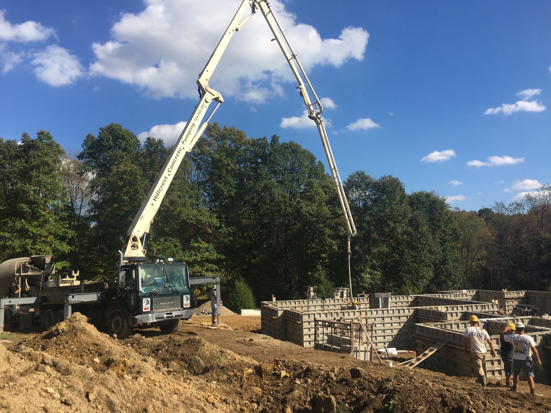 Concrete pump truck pouring concrete into forms on a construction site. Blue sky, green trees, workers present.