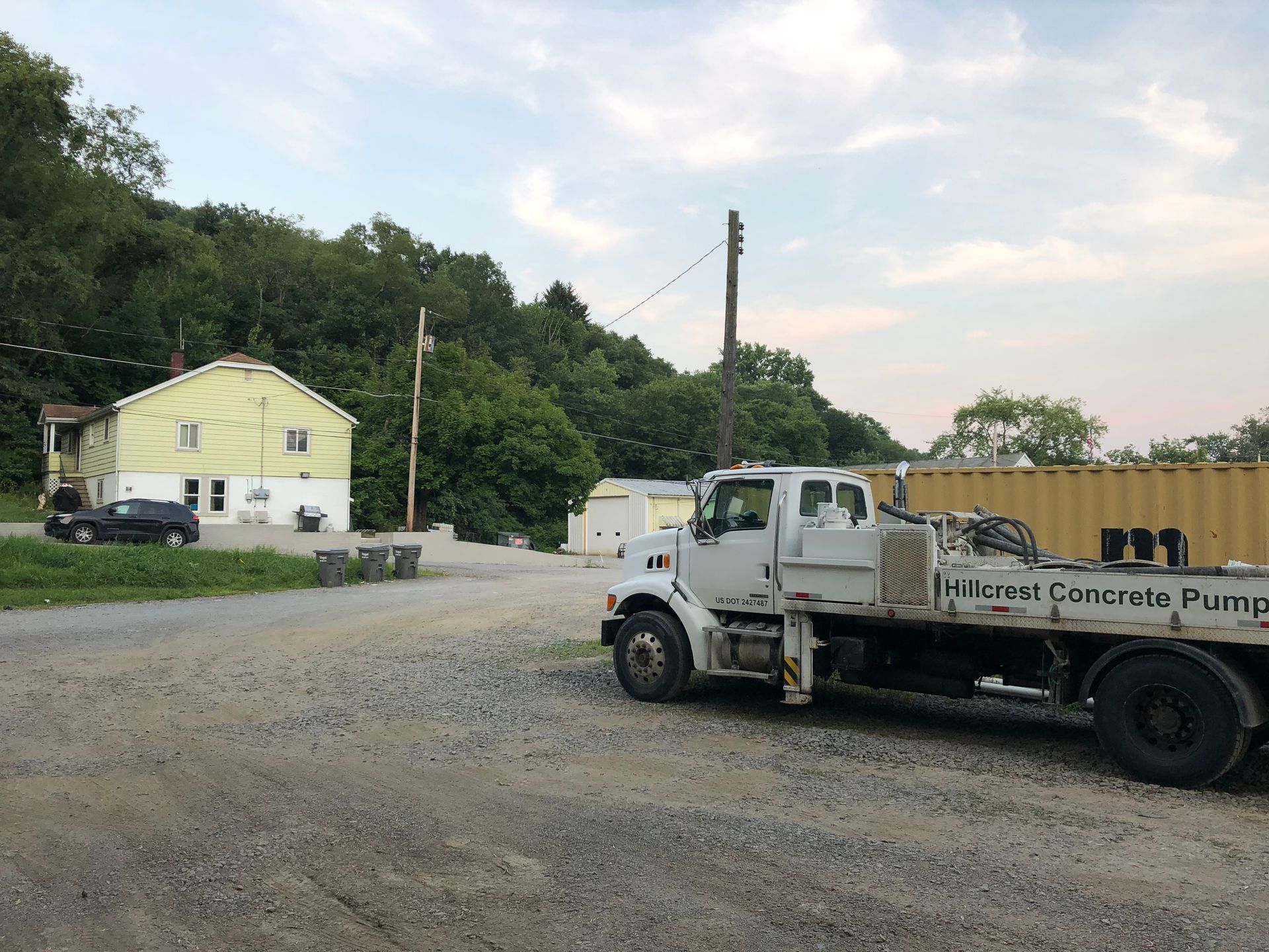 Gravel lot with a concrete pump truck, a pale yellow building, and trees on a hill under a cloudy sky.