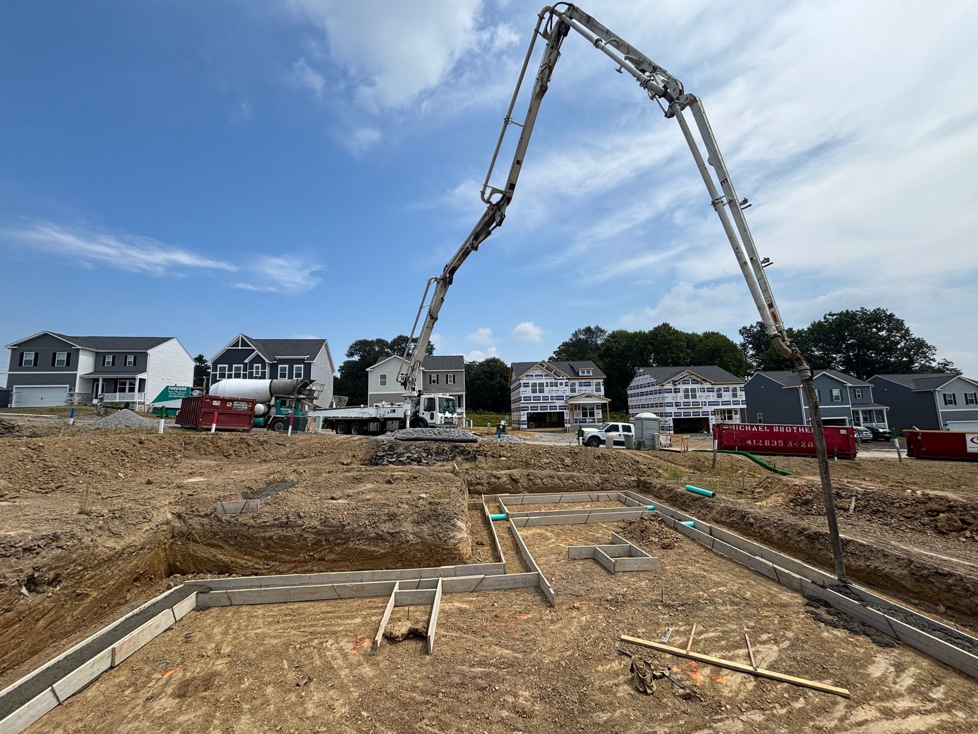 Construction site with concrete pump truck pouring concrete foundation. Houses in background, blue sky.
