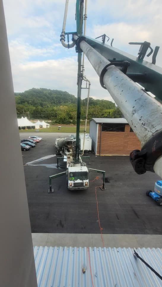 Concrete pump truck at work, parked on asphalt. Boom extended, with a building and wooded hills in the background.