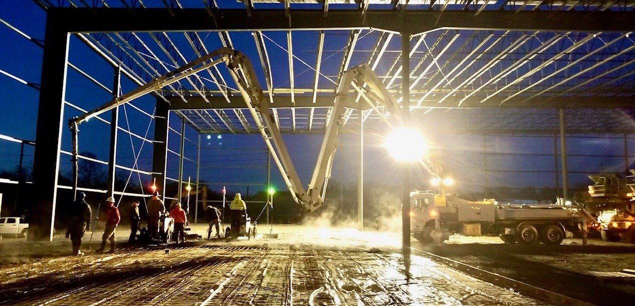 Construction workers pouring concrete at night using a boom truck, under a steel structure.