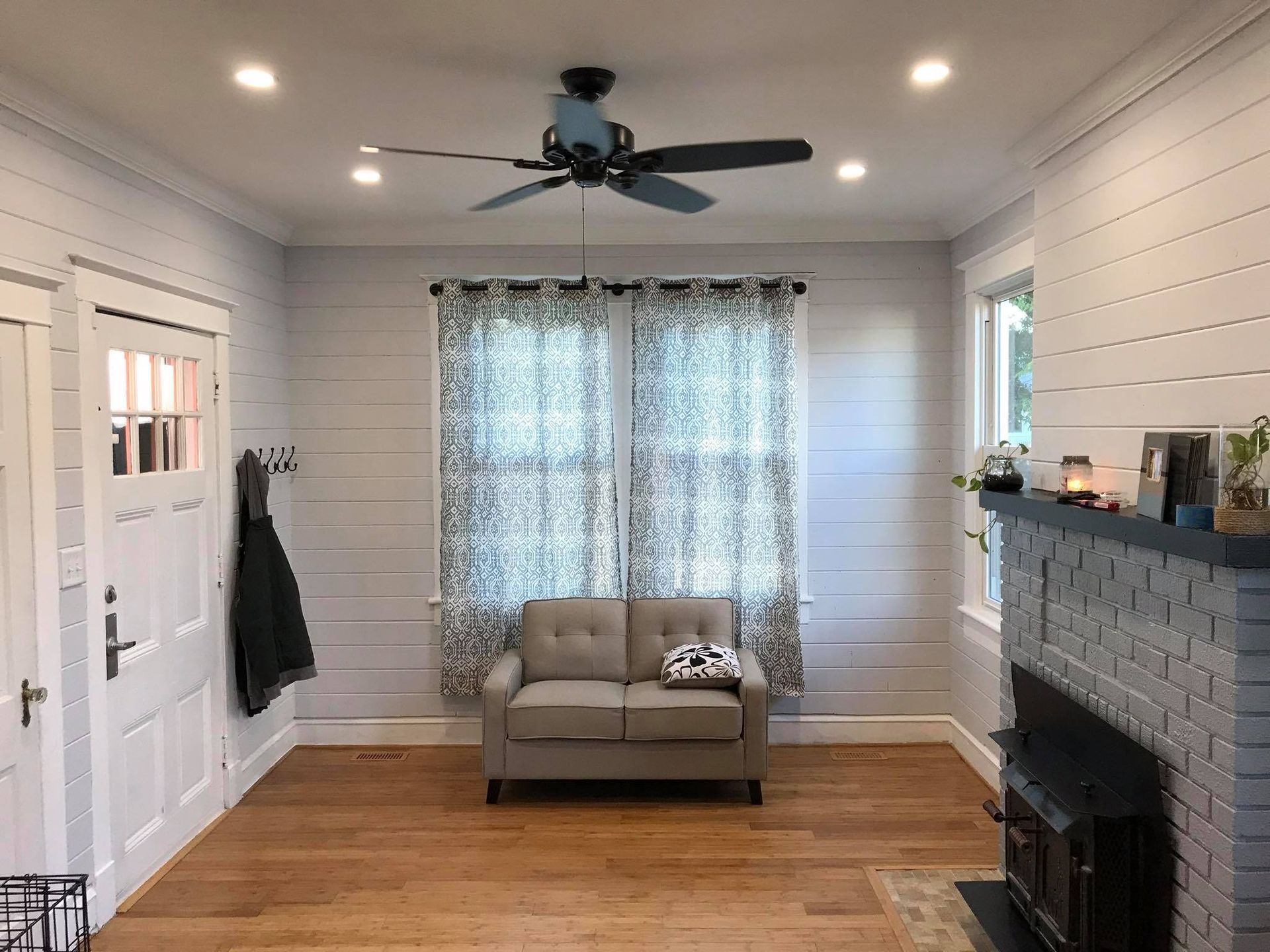 Bright living room with white shiplap walls, gray couch, fireplace, ceiling fan, and large curtained window.