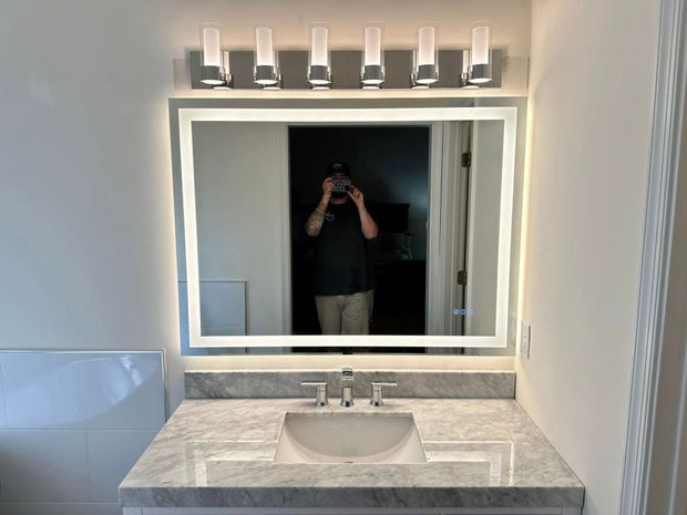 Bathroom vanity with large lit mirror, marble countertop, and a person reflected taking the photo.