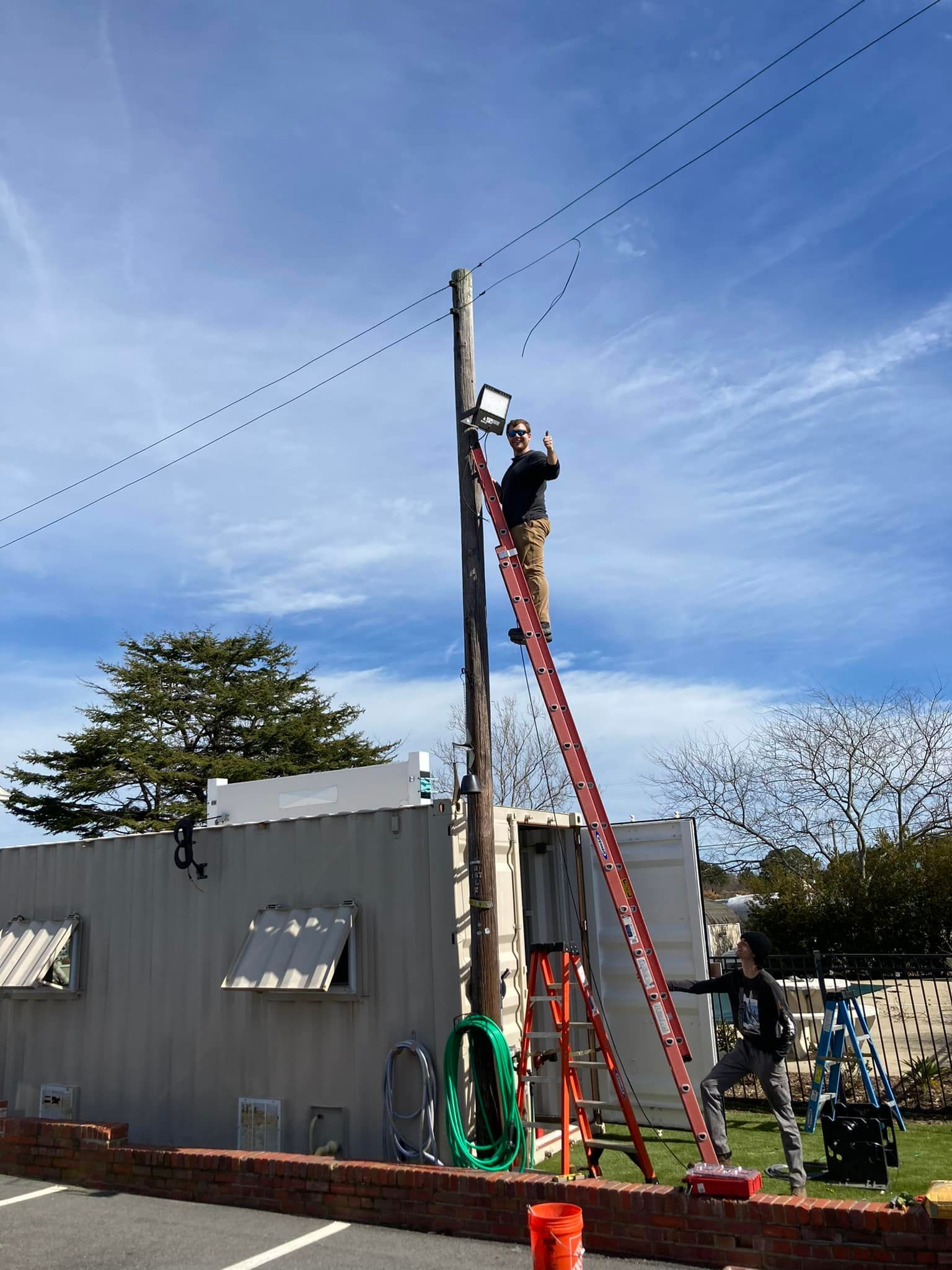 Worker on a ladder fixing a utility pole beside a gray building under a blue sky