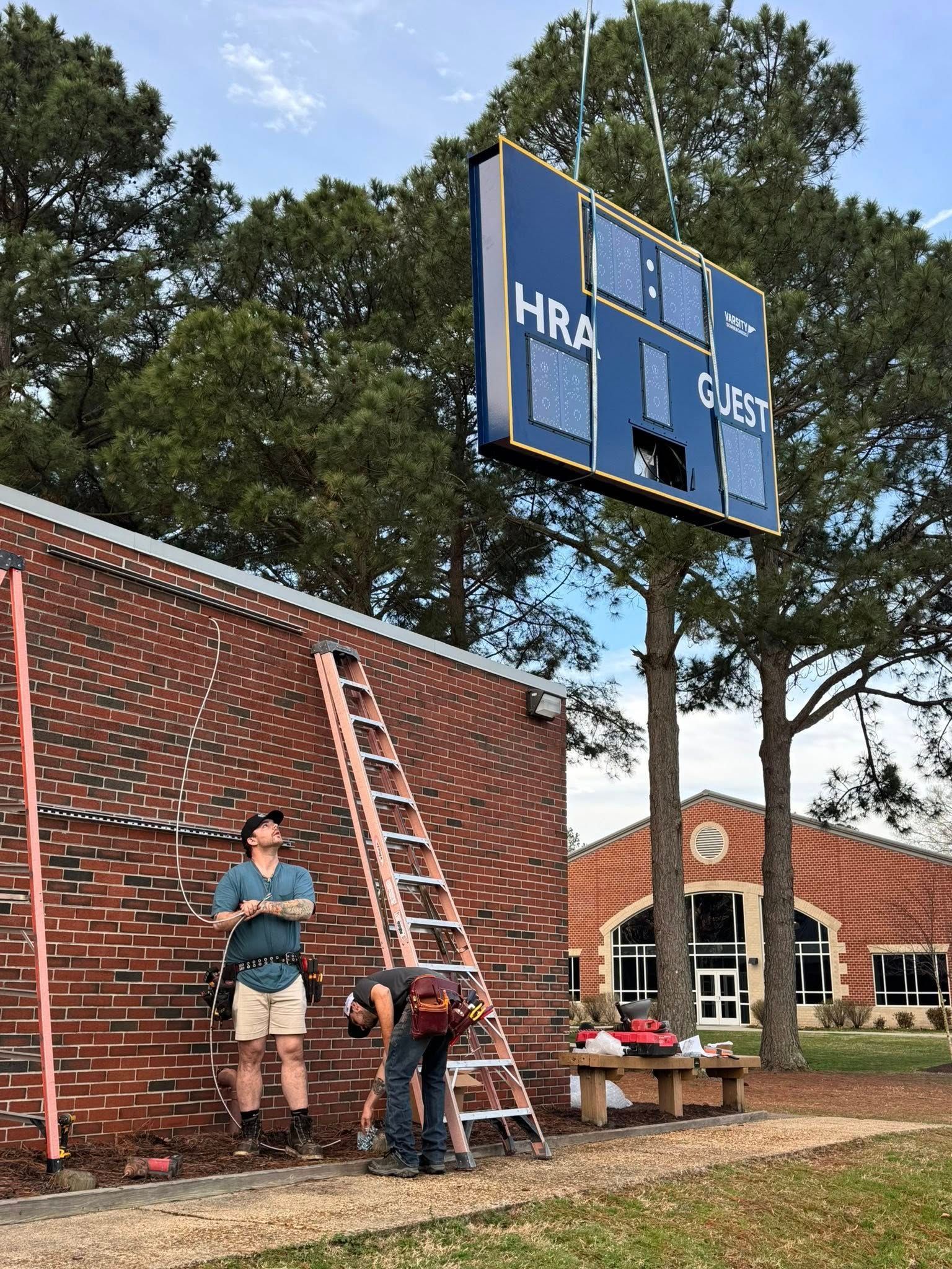 People on a ladder repairing a blue scoreboard beside a brick building and trees.