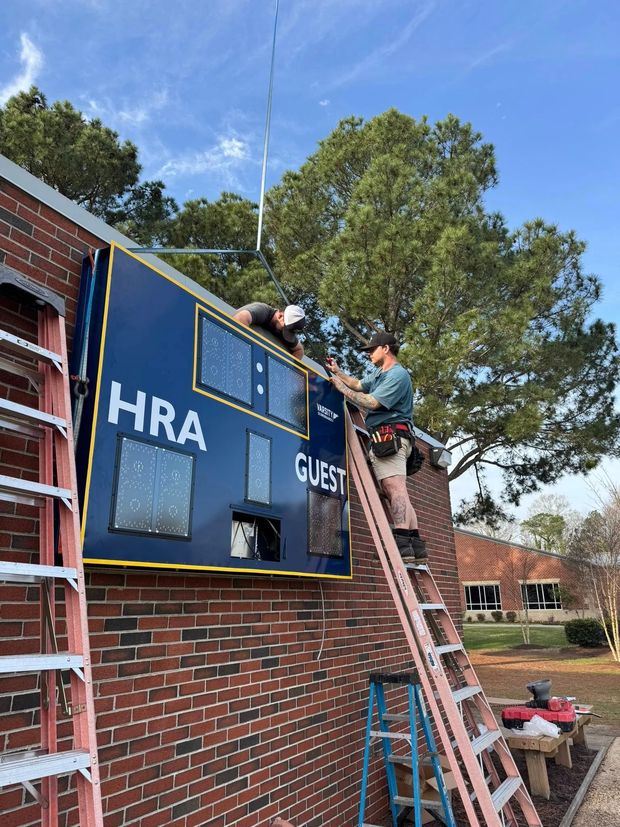 Workers installing a blue HRA sign on a brick building with a ladder outdoors