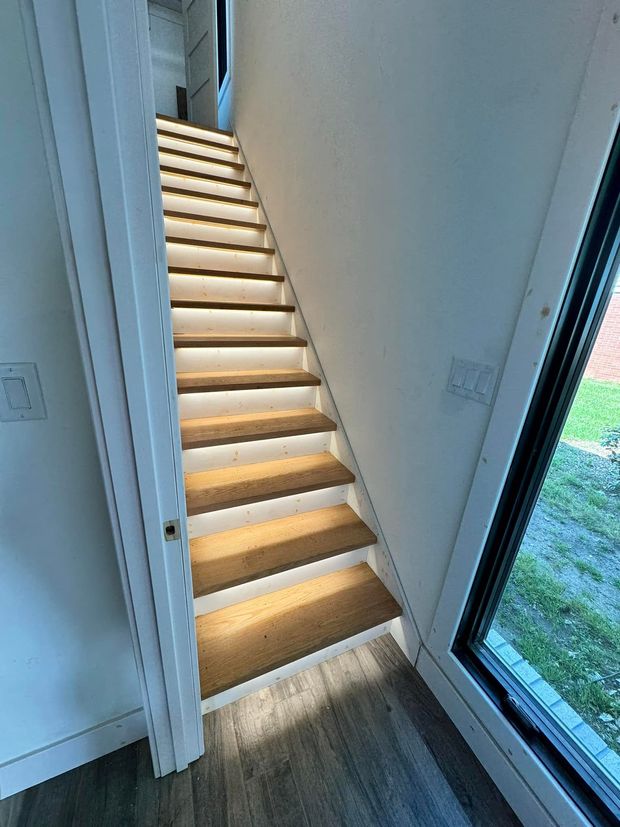 Narrow indoor staircase with illuminated wooden steps beside a tall window and white walls