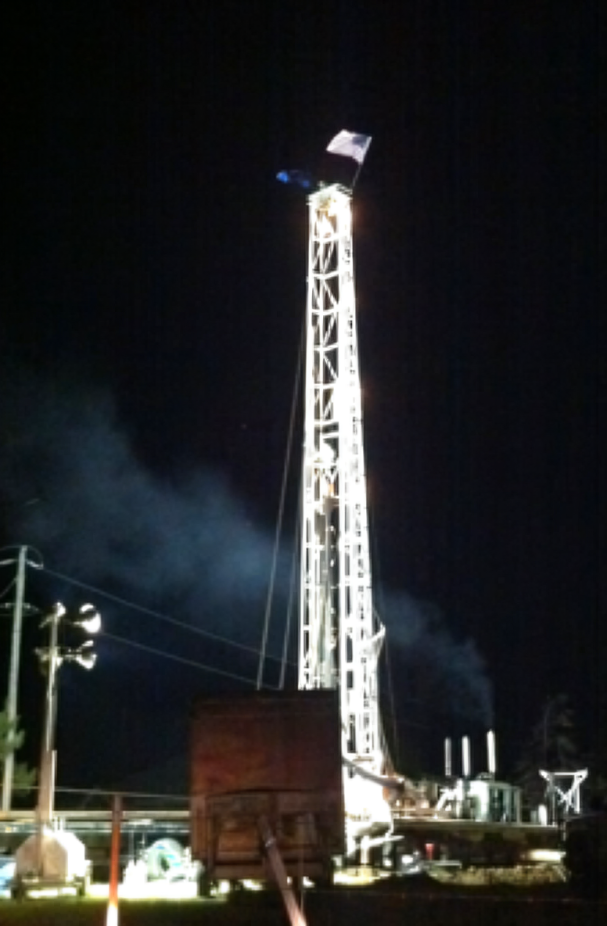 Nighttime oil drilling rig lit brightly against a dark sky, with machinery and power lines below