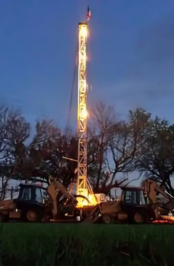 Illuminated drilling rig at dusk, with construction vehicles and bare trees in the background.
