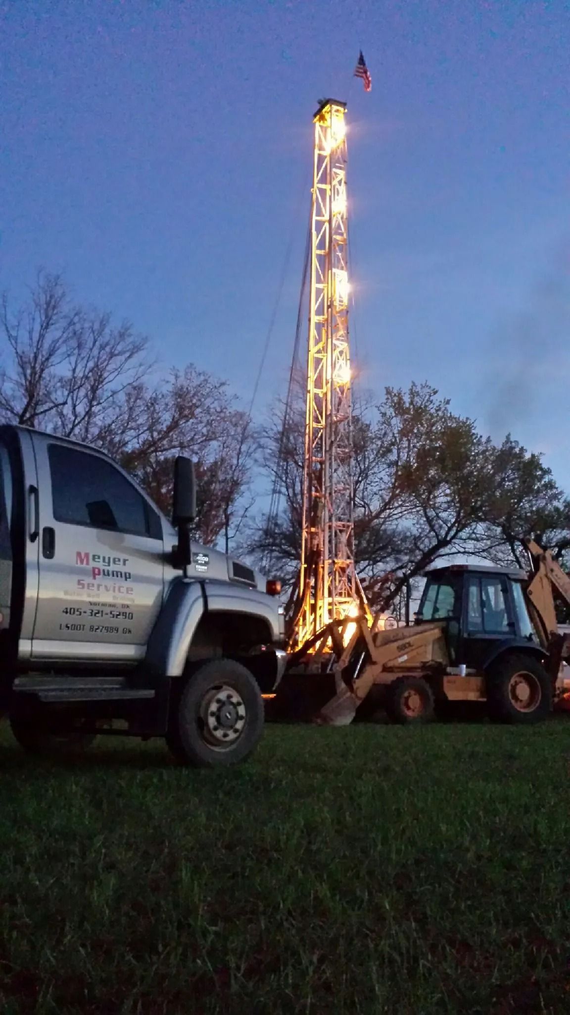 Drilling rig lit at dusk beside a white truck and skid steer on a grassy site
