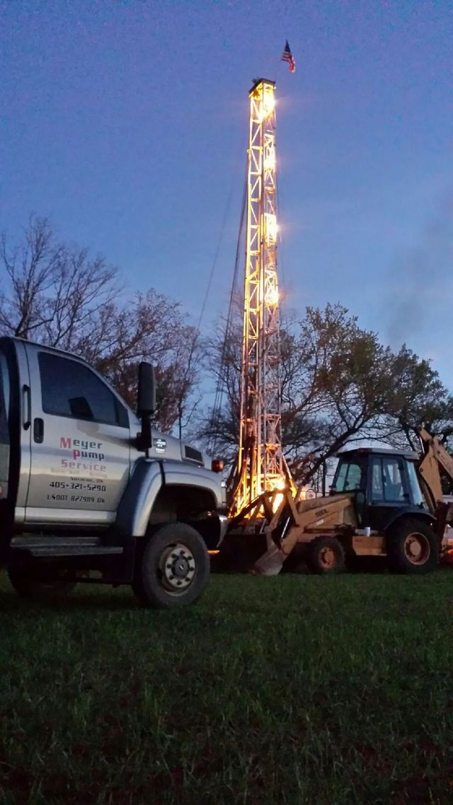 Drilling rig lit at dusk beside utility truck and excavator on a grassy field