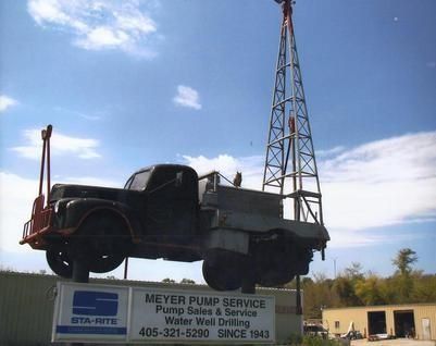 Old black truck on a Meyer Pump Service sign, with a tall metal rig against a blue sky.