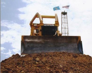 Bulldozer pushing a large mound of dirt under a cloudy sky, with flags on the machine.