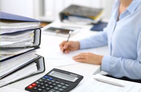 Woman at desk writing, with calculator and stacked files.