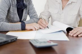 Two people reviewing documents, one signing a paper, on a table with a laptop and calculator.