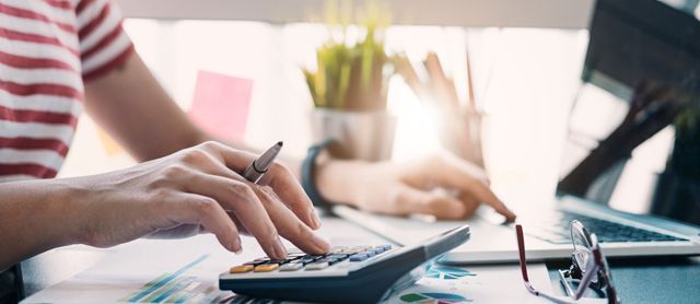 Person using a calculator and laptop, working at a desk, sunlight in background.