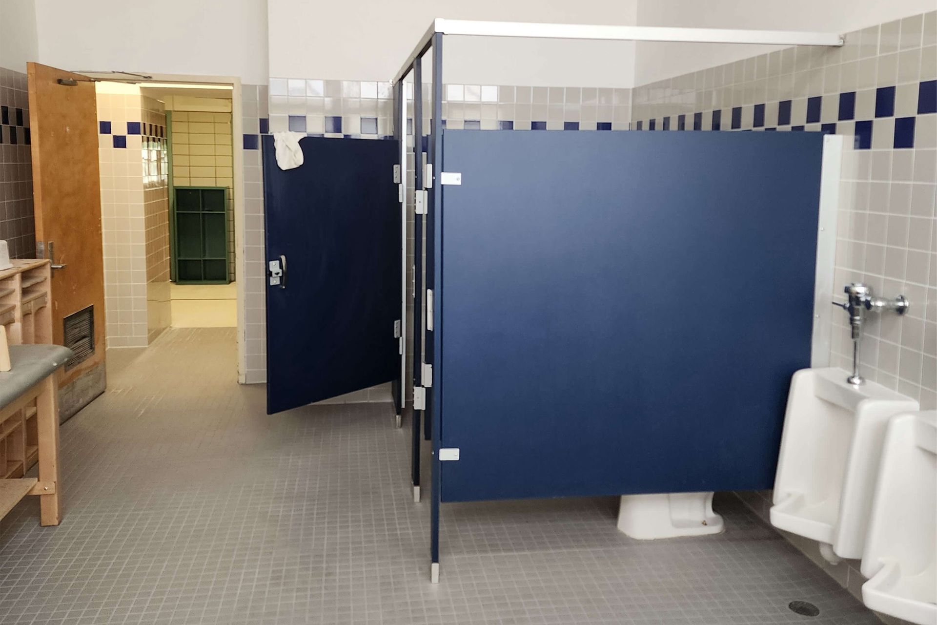 Public restroom with blue stalls and urinals. Tile floor and walls. Open door to hallway.
