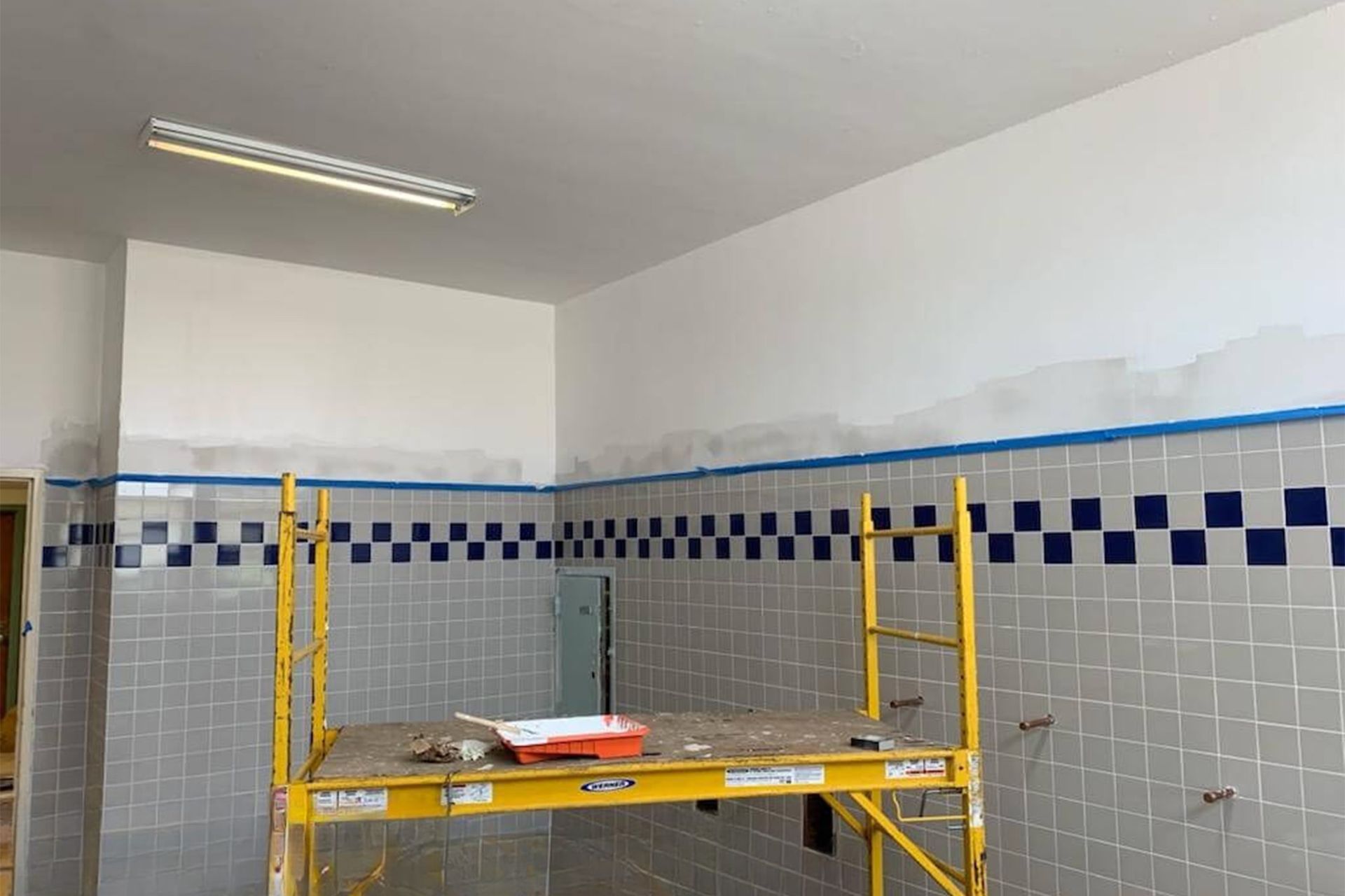 Room with gray tiled walls, blue checkered border, and white upper walls being painted. Yellow scaffold present.