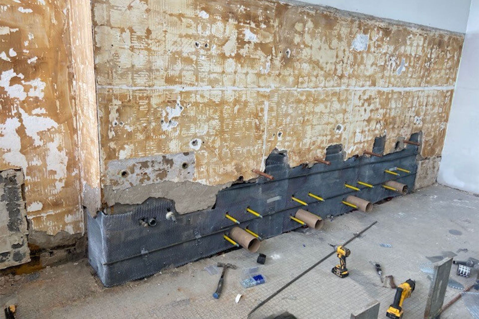Wall construction with exposed insulation and pipes. Tools and debris on the floor.