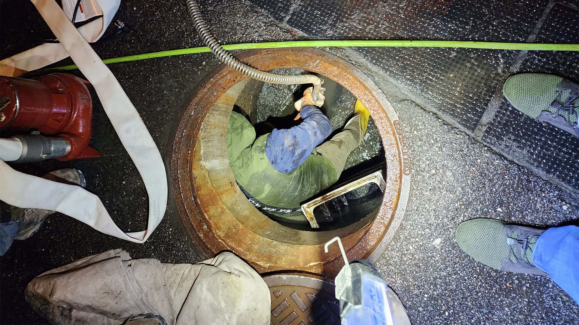 Person inside an open manhole, surrounded by tools and equipment. Dark setting with various cords and materials visible.