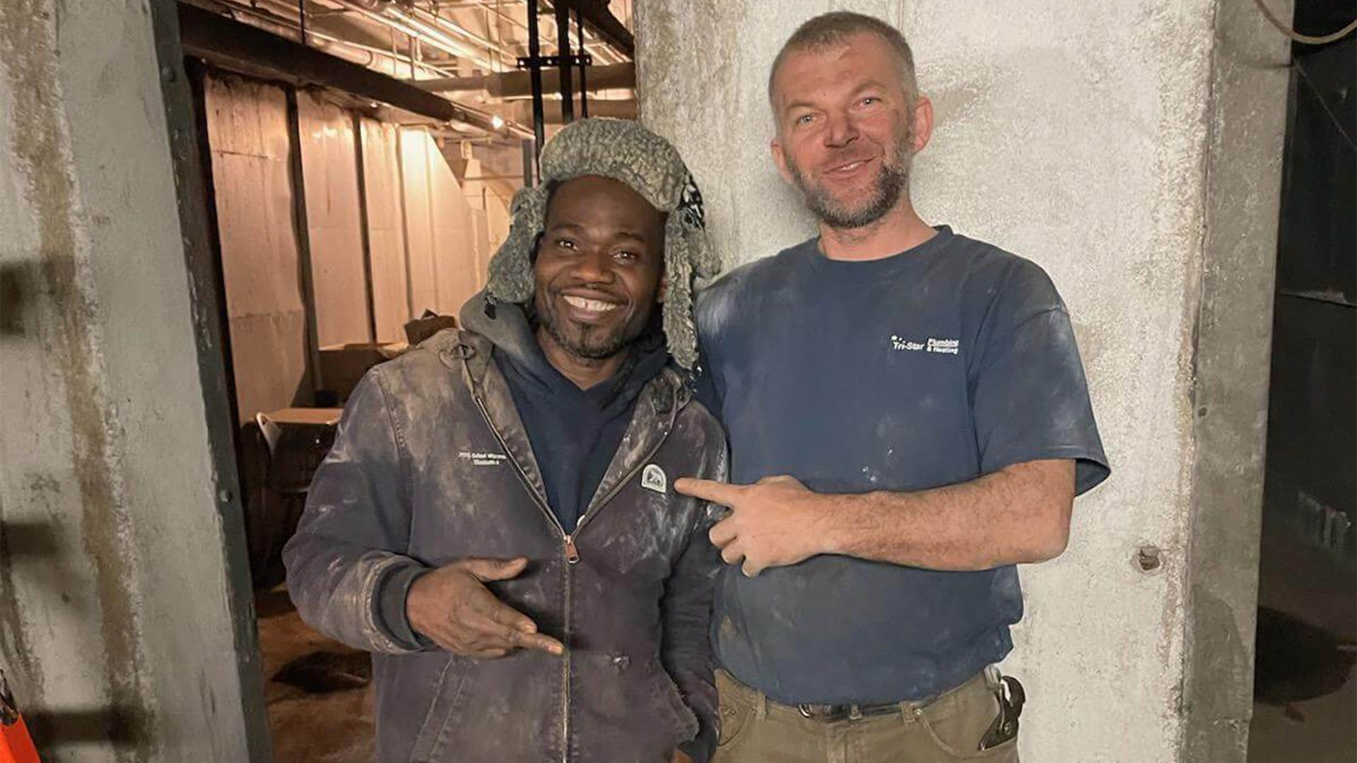 Two construction workers smiling indoors, one pointing. Dark clothing, concrete walls.