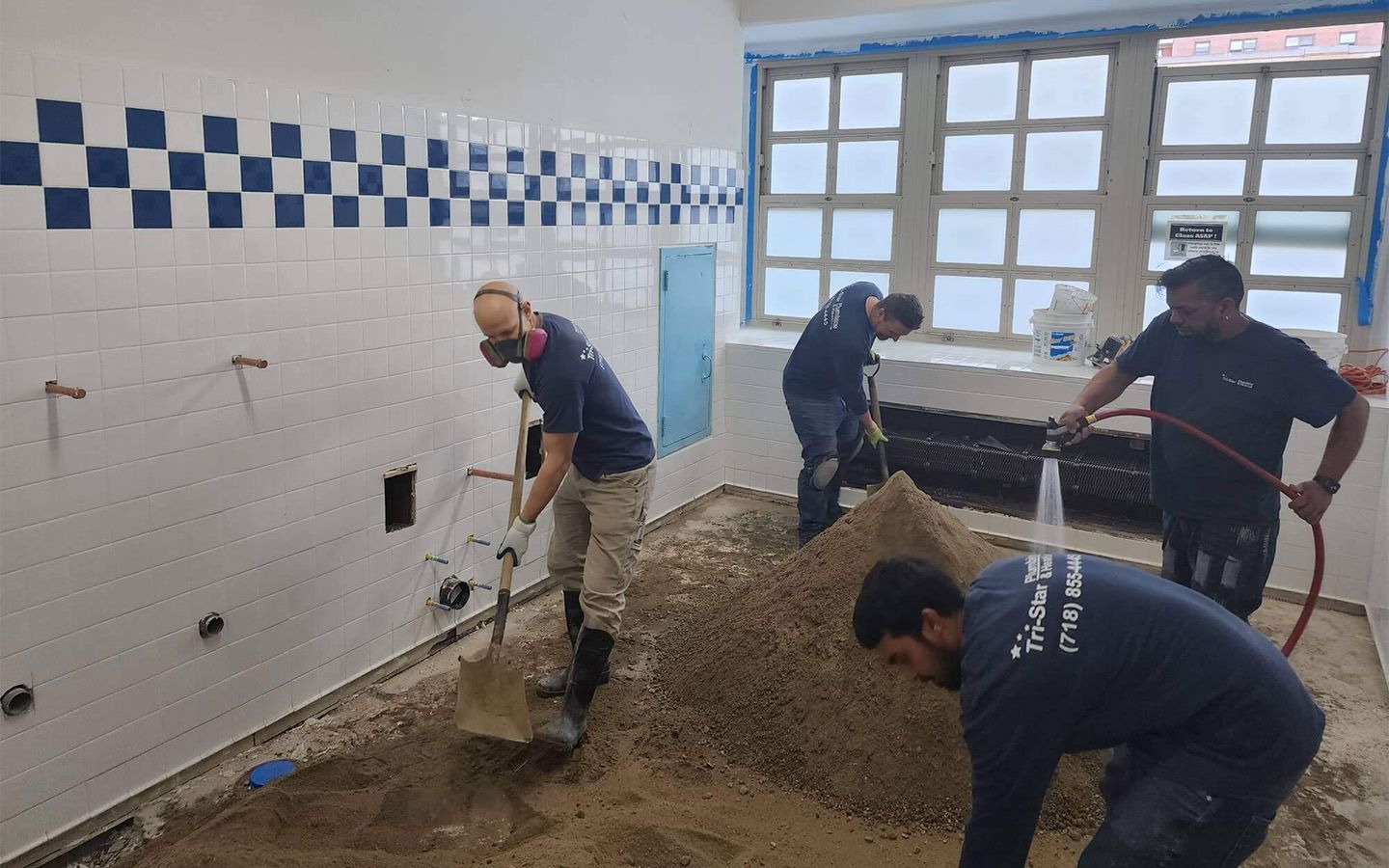 Workers renovating a room with tiled walls and a window. One shovels sand, another hoses it down.
