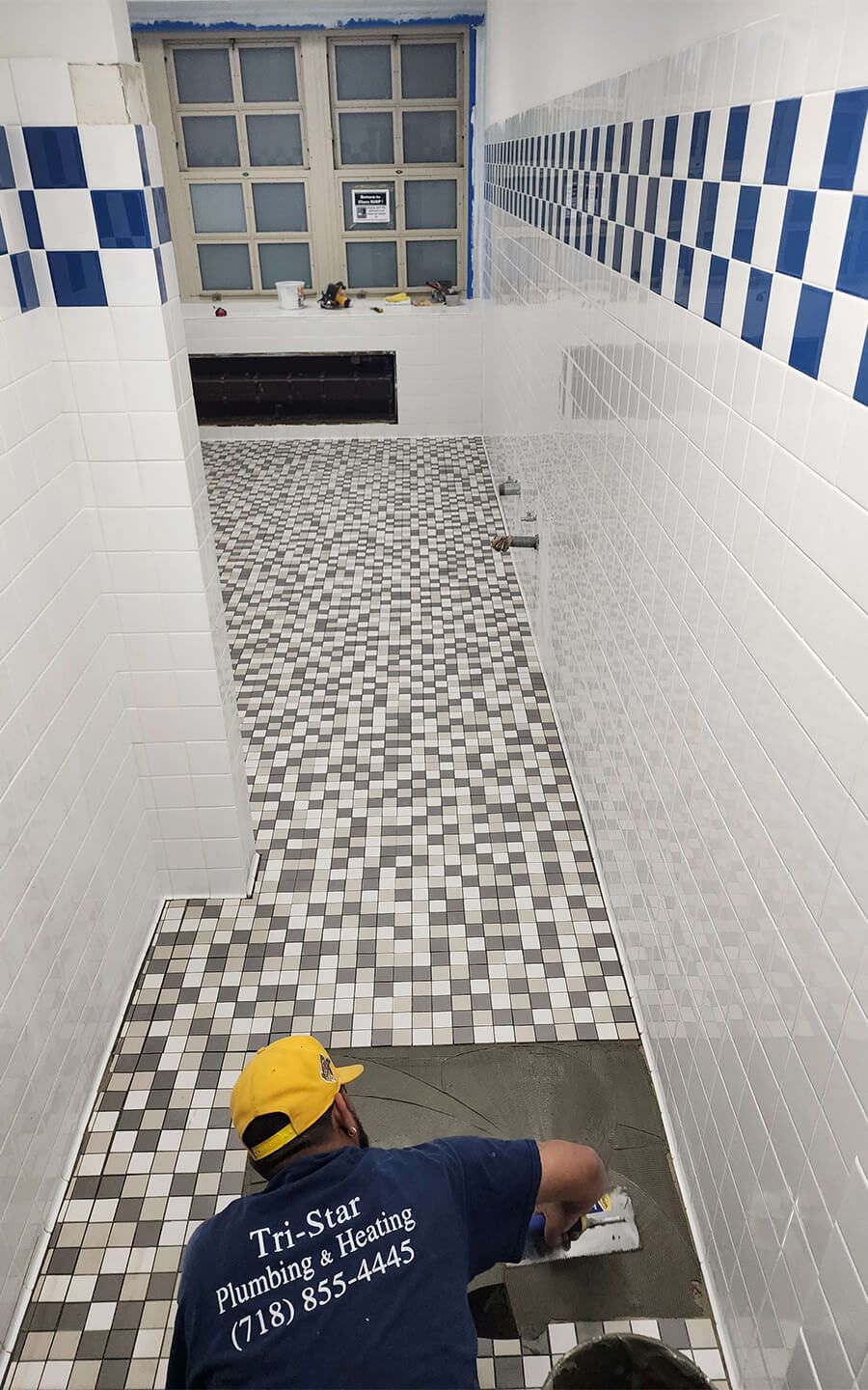 Person tiling a bathroom floor with multi-colored mosaic tiles, walls are white and blue checkered.
