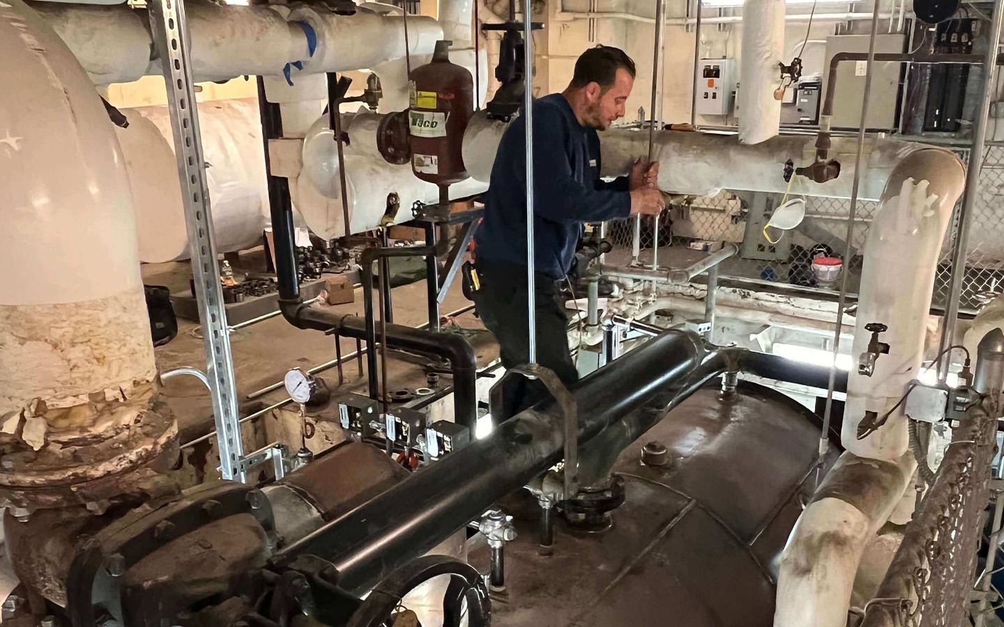 Man in blue shirt works on pipes in a mechanical room, surrounded by equipment and insulation.