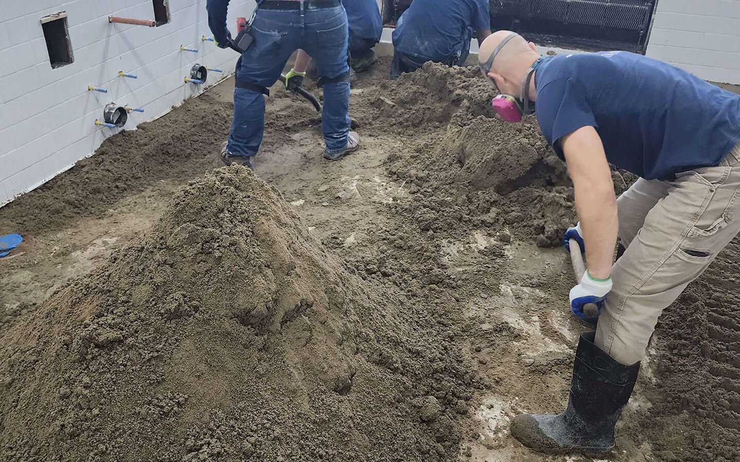 Construction workers shoveling loose material over a concrete foundation; one wears a respirator.