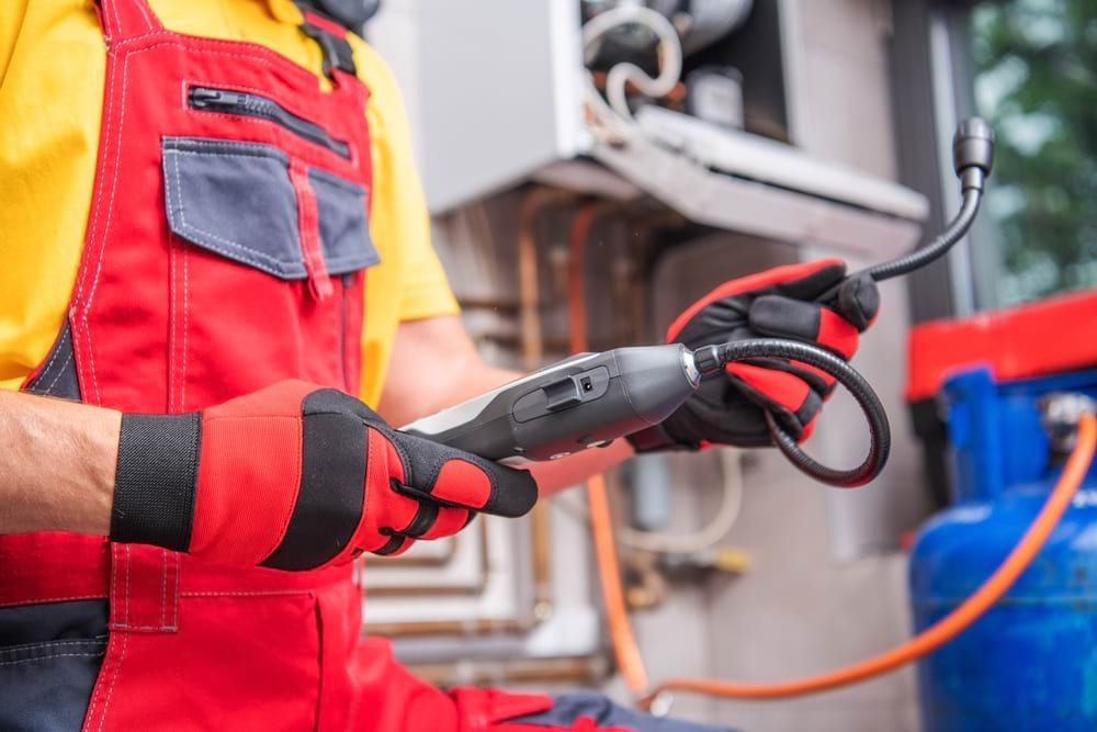 Plumber in red overalls holding a tool, inspecting gas line near a blue propane tank.
