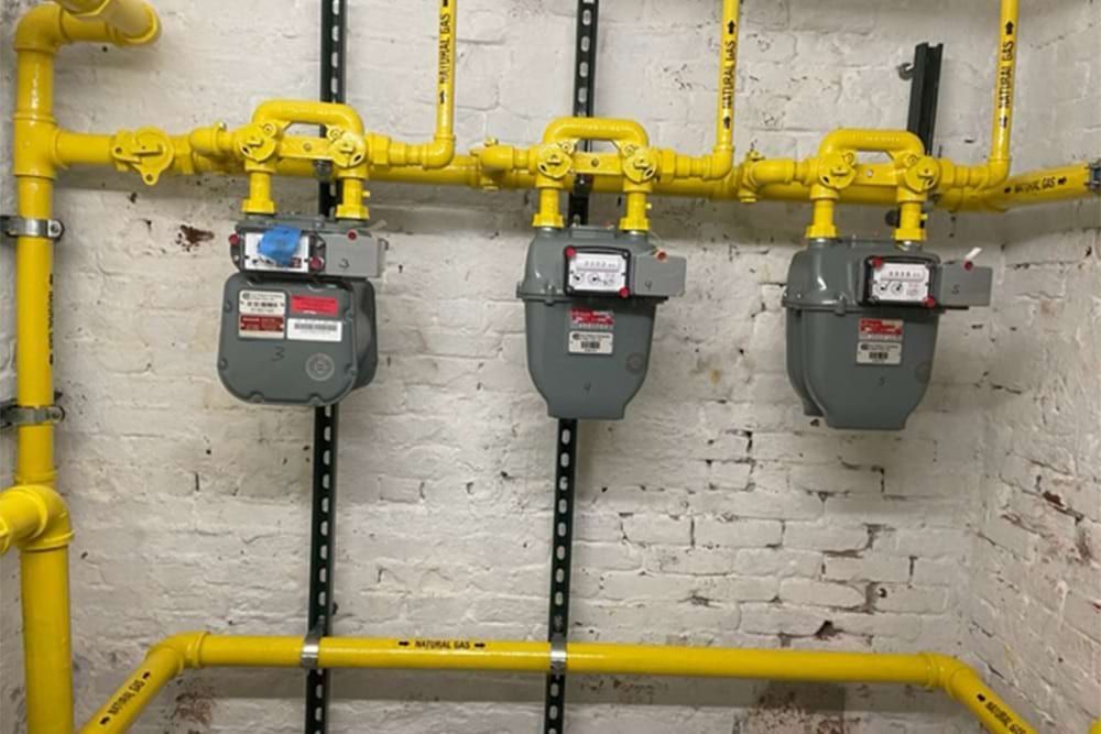 Three gray gas meters with yellow pipes against a brick wall.
